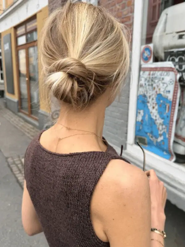A blonde woman in a low messy bun stands on a city sidewalk, holding sunglasses. She wears a sleeveless brown knit top made from the PetiteKnit Bea Top neuleohje, ENG, accessorized with delicate gold necklaces. Storefronts are behind her.