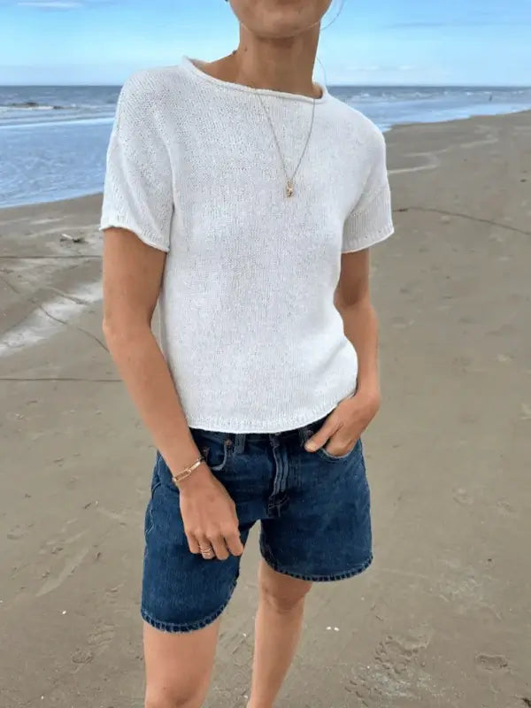 Someone wearing a white PetiteKnit Cloud tee neuleohje and denim shorts stands on a sandy beach with the ocean behind them, one hand in their pocket and the other at their side.