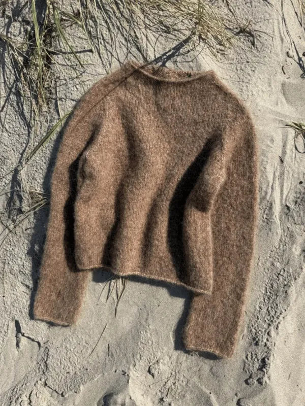 The Petiteknit IVY SWEATER neuleohje rests on pale sand with dry grass, creating a cozy, natural scene—ideal inspiration for your next knitting project.