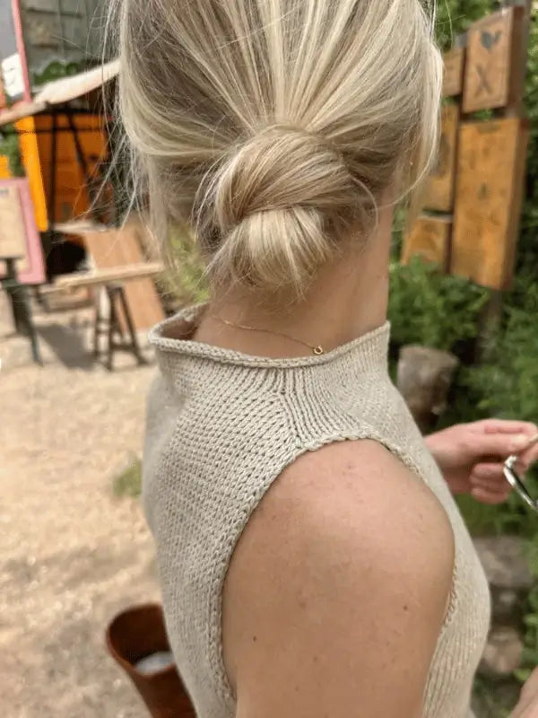 A blonde woman with a low bun stands outdoors facing away, holding sunglasses. She’s wearing the PetitKnit Ivy Top Neuleohje, surrounded by greenery and wooden signs in the background.