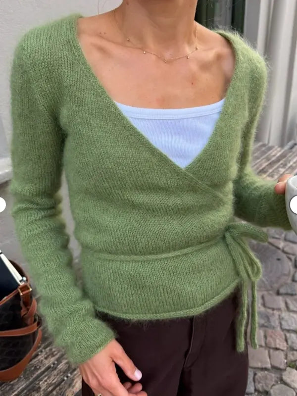 A person wears a soft green sweater—the PetiteKnit Lucy Wrap, known for its tie at the waist—over a white top and dark pants. Part of a bag and a stone street are visible in the background, making this look inspired by the English neuleohje both cozy and stylish.