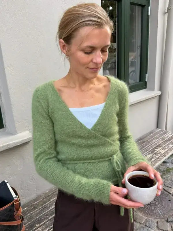 Wearing a PetiteKnit Lucy Wrap - English neuleohje in light green, a woman stands outside by a bench, cradling a cup of coffee and looking down at her drink.