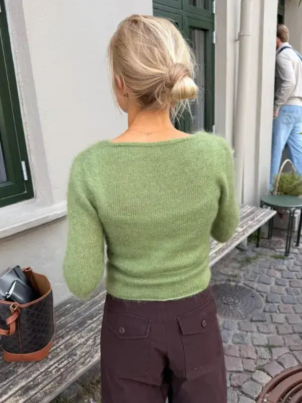 A woman with blonde hair in a low bun, wearing a fuzzy green PetiteKnit Lucy Wrap sweater and dark pants, stands with her back to the camera on a cobblestone sidewalk near a bench and a green window.