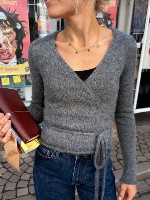 Lucy, dressed in a gray PetiteKnit Lucy Wrap—English neuleohje—and jeans, holds a book and a paper bag. She stands on a cobblestone street adorned with colorful posters, creating the perfect backdrop to highlight this effortlessly stylish knitwear look.