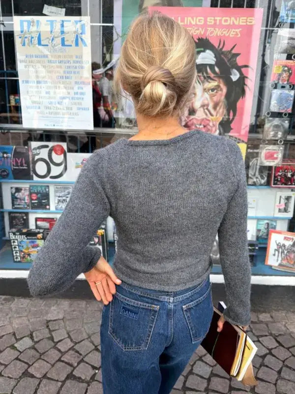 A woman with blonde hair in a low bun—possibly Lucy—stands facing a shop window adorned with music posters and albums. She wears a gray sweater made from the PetiteKnit Lucy Wrap (English knitting pattern), paired with blue jeans, and holds a notebook and phone behind her back.