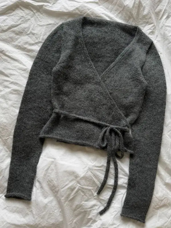 A PetiteKnit Lucy Wrap (English neuleohje) in dark gray, featuring long sleeves and a tie belt, is laid flat on a wrinkled white fabric background.