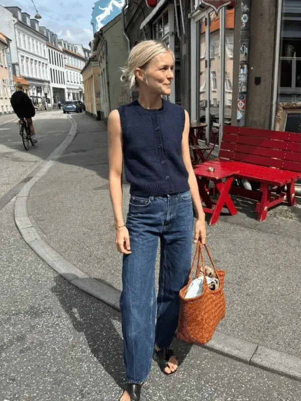 A woman wearing a navy sleeveless top and blue jeans—styled after the PetiteKnit Maggie Vest neuleohje—stands on a city street with a woven tote bag. Red benches, buildings, and a cyclist complete the scene.