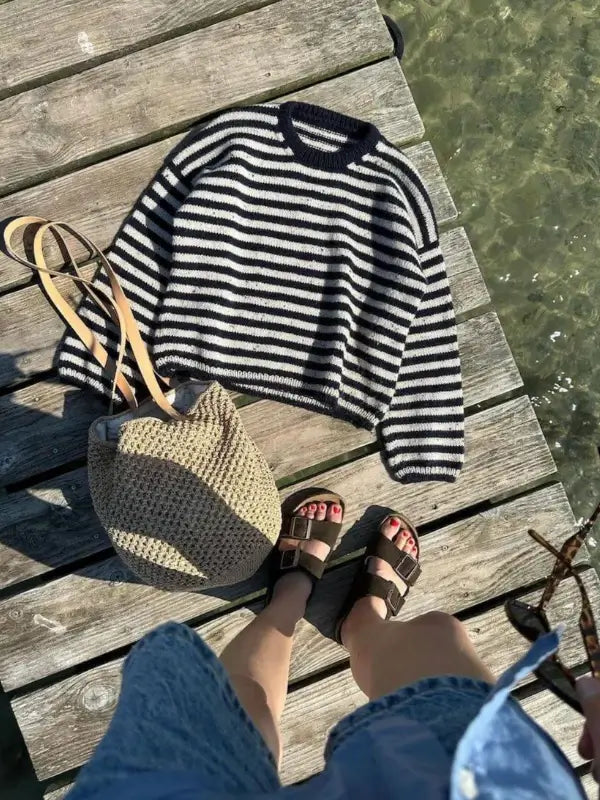 A person stands on a wooden dock above clear water, dressed in sandals and a blue dress. Beside them lies a black and white striped sweater inspired by the PetiteKnit Olga sweater neuleohje, along with a woven bag featuring tan straps. A PetiteKnit Olga sweater neuleohje is gently placed next to the bag, suggesting peaceful knitting sessions by the water.