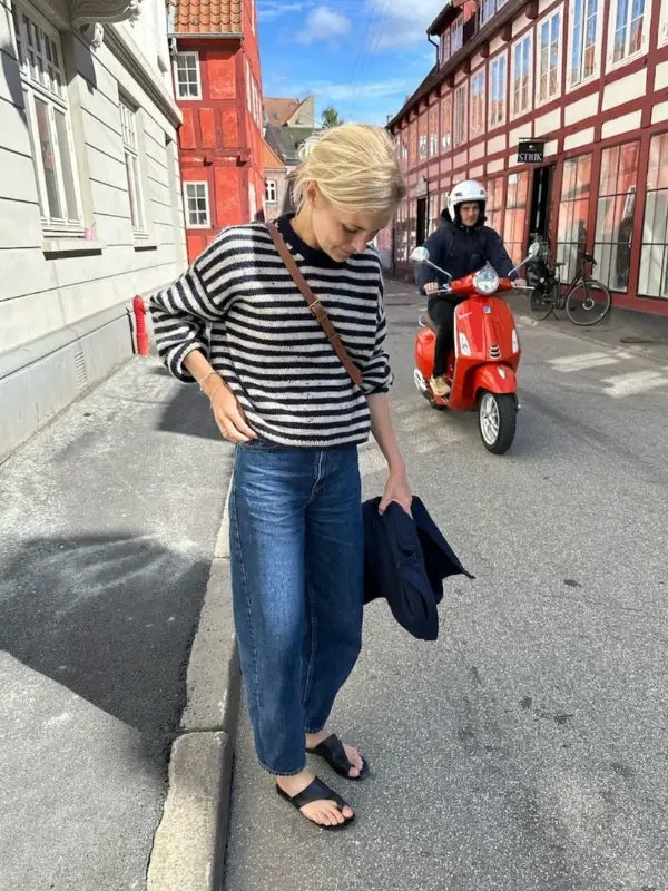 A woman wearing a striped sweater—likely crafted from the PetiteKnit Olga sweater neuleohje—paired with jeans and sandals stands on a street holding a jacket. Behind her, someone rides a red scooter. The street, lined with colorful buildings, creates a lively backdrop reminiscent of any scene found in neuleohje suomeksi.