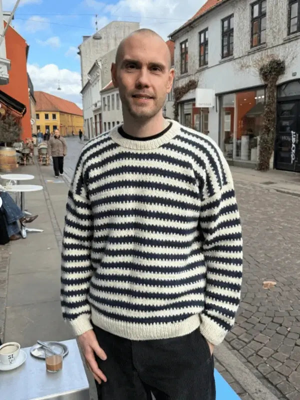 A smiling man with a shaved head stands outdoors on a cobblestone street, wearing a PetiteKnit Otto Sweater neuleohje. Cafés and shops line the street, with cups on a nearby table under the partly cloudy sky.