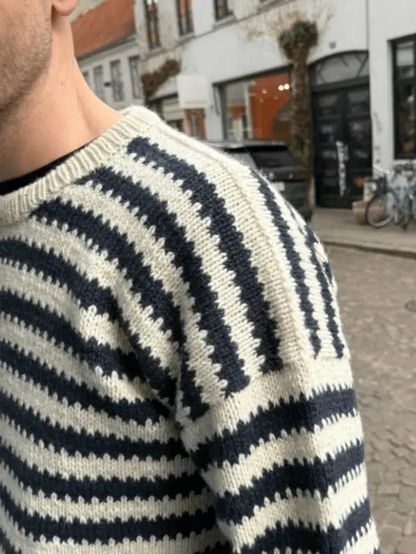 A close-up of a person wearing a cream and navy blue striped PetiteKnit Otto Sweater neuleohje, standing outdoors on a cobblestone street with buildings, parked cars, and bicycles in the background.