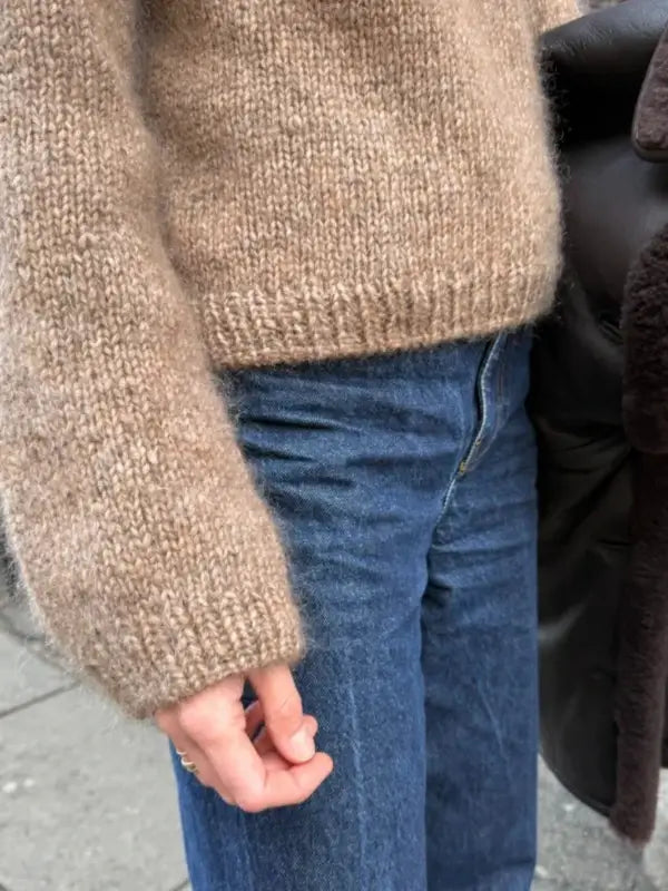 A person wearing a brown knitted sweater made from the PetiteKnit Sunday Sweater neuleohje, Suomi, paired with blue jeans stands outside, holding a dark brown coat in their left arm.