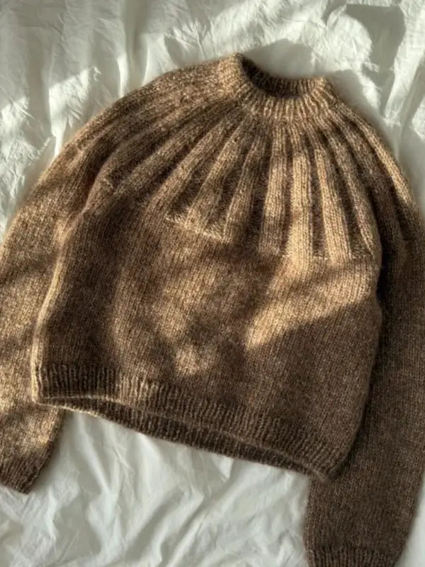 A PetiteKnit Sunday Sweater neuleohje, Suomi—a brown, knitted sweater with ribbed cuffs and neckline—lies flat against a white, wrinkled fabric background. Sunlight gently casts soft shadows over the cozy design.