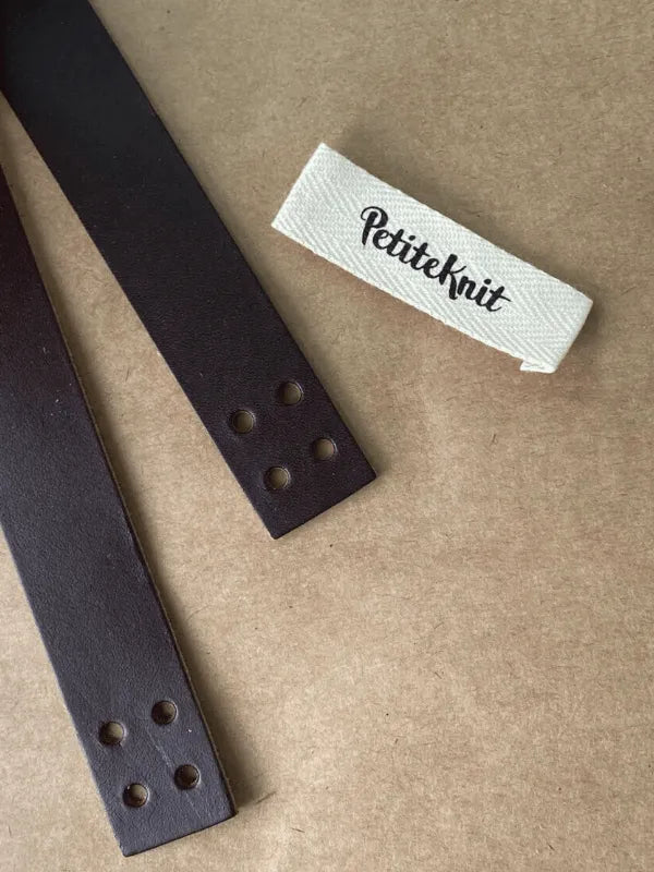 Two dark brown PETITEKNIT Nahkahihnat Florence laukkuun ruskeat, each with four holes at the ends, are on a brown surface beside a white PetiteKnit label—ideal for finishing your Florence laukku project.