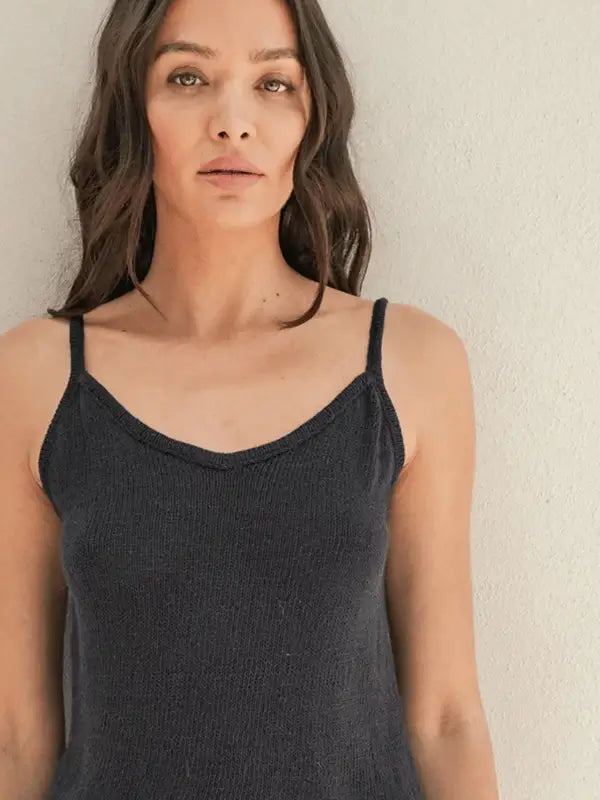 A woman with long brown hair wears the Sandnes Garn Clementine Top, knit from Tynn Line yarn, standing against a light wall and looking directly at the camera with a neutral expression.