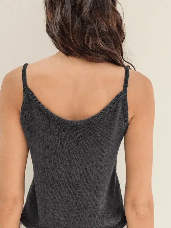 A woman with long, wavy brown hair is seen from the back wearing the Sandnes Garn Clementine Top, made using Tynn Line yarn. The dark gray knit top features thin straps and a lovely pattern, displayed against a plain light background.
