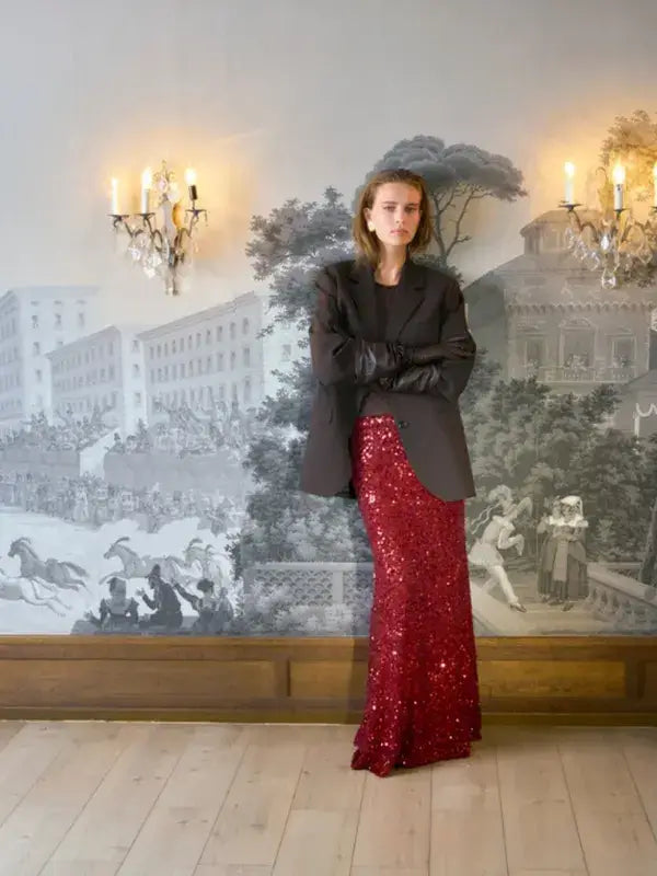 A person wears a dark blazer, a dark top, and a floor-length Sandnes Garn Vea Skirt – neulesetti Paljett covered in sequined yarn.