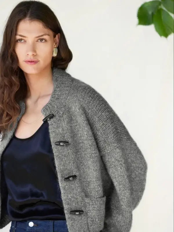 A woman with long brown hair wears a gray knit bomber jacket created from the Sandnes Garn 2606-08 DIY Bomber Jacket – neuleohje Suomi, PDF pattern over a black satin top, standing against a light background with a green leaf visible in the top right corner.