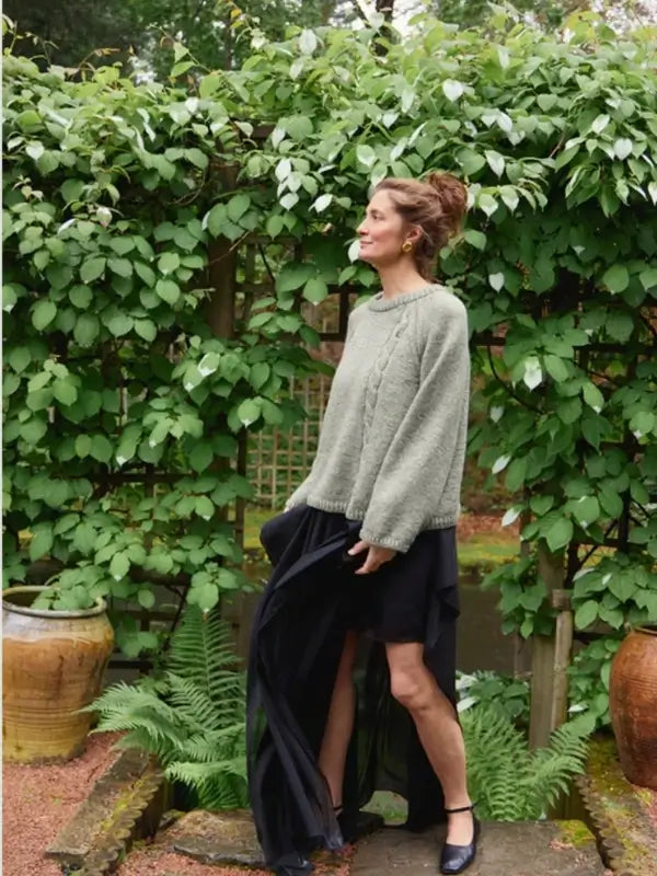 A woman wearing the Sandnes Garn 2606-10 DIY Laura Sweater – neuleohje suomeksi, PDF in grey and a black skirt stands outdoors beside green leafy bushes and large pots, one hand holding her skirt and her hair styled up.