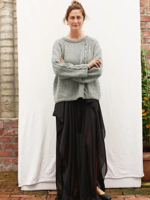 A person stands in front of a white backdrop wearing a gray Sandnes Garn 2606-10 DIY Laura Sweater – neuleohje suomeksi, PDF with cable knit detail and a long, flowing black skirt. Brick walls and plants with yellow flowers are visible in the background.