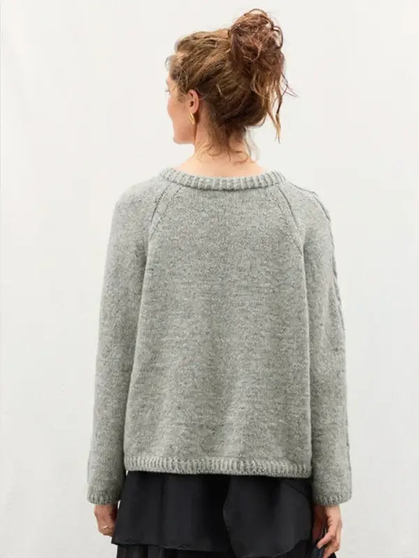 A person with curly hair styled in a messy bun wears a loose, light gray knit sweater—similar to the Sandnes Garn 2606-10 DIY Laura Sweater (neuleohje suomeksi, PDF)—paired with a black skirt, standing with their back to the camera against a plain light background.