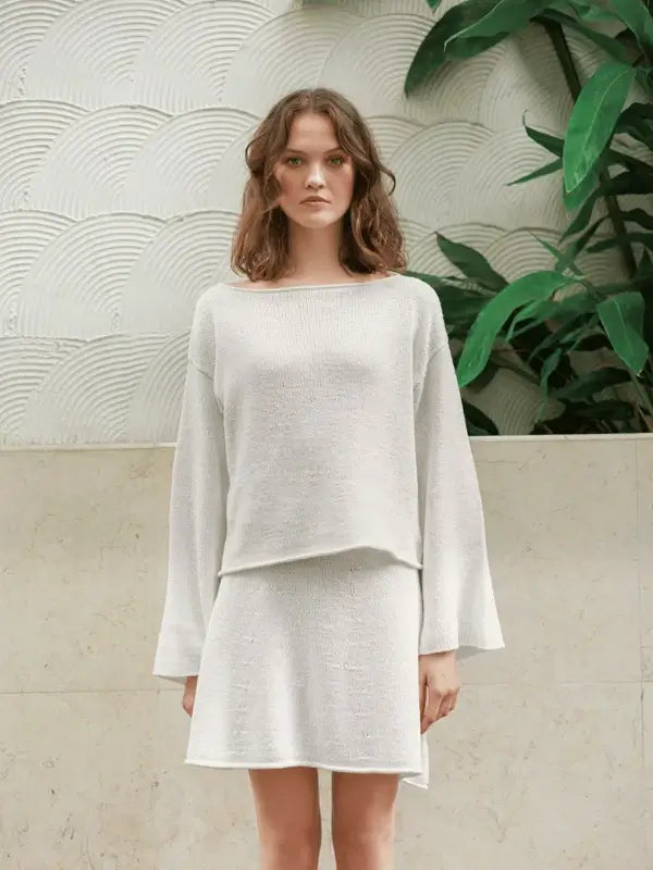 A woman stands indoors before a white textured wall and green plants, wearing a loose Milly Sweater & Skirt Neuleohje Suomi made from Sandnes Garn line yarn. She gazes straight ahead with a neutral expression.