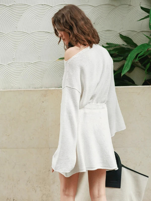 A woman with wavy brown hair wears a loose white sweater dress inspired by the Milly Sweater & Skirt Neuleohje Suomi + Sandnes Garn line, standing sideways against a textured white wall with greenery and a beige surface.