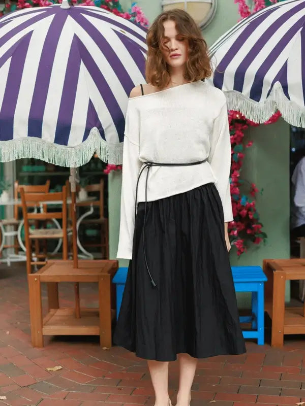 A woman with wavy hair stands outdoors under a purple and white striped umbrella, wearing the Milly Sweater & Skirt Neuleohje Suomi made with Sandnes Garn Line. Pink flowers and wooden chairs are in the background.