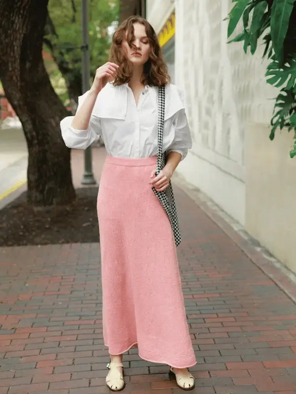A woman stands on a brick sidewalk wearing the Milly Sweater & Skirt Neuleohje Suomi made with Sandnes Garn Line, paired with cream shoes and a checkered bag. Green trees and a cream wall are in the background.