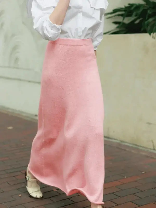 A person in a Milly Sweater & Skirt Neuleohje Suomi +Sandnes Garn line outfit—a white long-sleeve blouse and high-waisted pink skirt—walks on a brick path, framed by light walls and green plants.