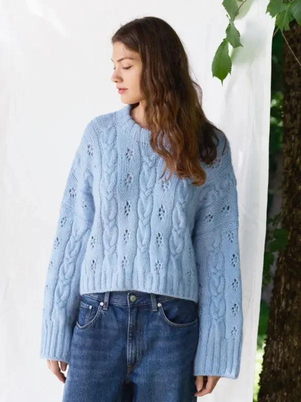 A woman with wavy brown hair wears a light blue cable-knit sweater—similar to the Sandnes Garn 2606-11 Junia Sweater (neuleohje suomeksi PDF)—paired with blue jeans, standing against a white backdrop with green leaves and a tree visible to the right.