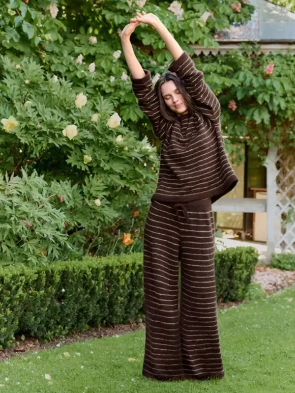 A person wearing the Sandnes Garn Riley Sweater & Riley Pants – neuleohje suomeksi, PDF set in brown and white stripes stands on grass, stretching with arms overhead amidst lush greenery and blooming bushes.