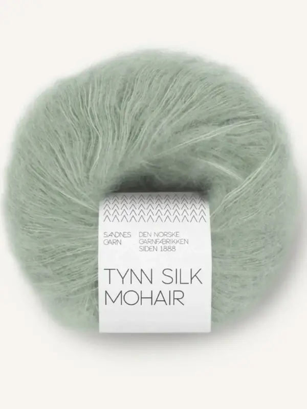 A ball of soft, pale green mohair-silk yarn with a white label reading “Sandnes Garn Tynn Silk Mohair” and Norwegian text above it, this luxurious thread comes from the renowned Sandnes Garn collection. Perfect for delicate projects, Sandnes Garn Tynn Silk Mohair offers an elegant sheen and a gentle touch.