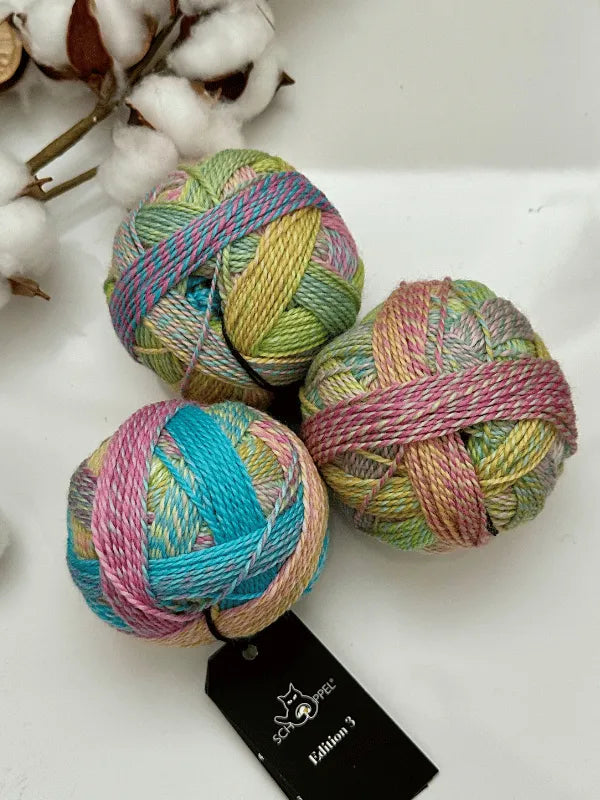 Three pastel-colored balls of Schoppel-Wolle Edition 3 yarn in green, blue, pink, and yellow rest on a white surface. One ball has a black product tag attached. Cotton stems are blurred in the background.