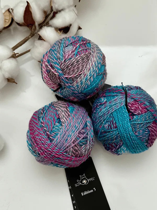 Three colorful balls of Schoppel-Wolle Edition 3 yarn in pink, purple, gray, and teal gradient rest on a white surface beside cotton stems. Below the yarn is a black ruler featuring the Schoppel logo and Edition 3 branding.