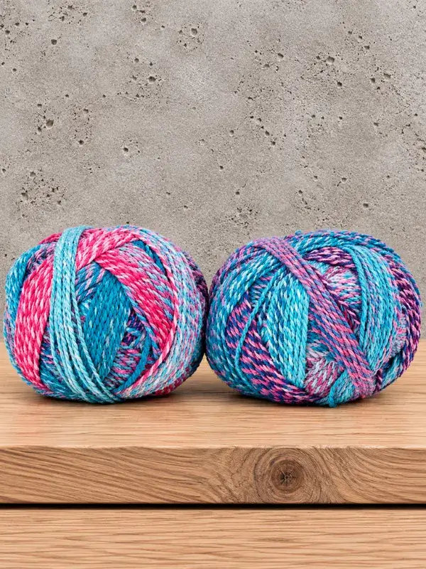 Two balls of Schoppel-Wolle Edition 3 variegated yarn in shades of pink, turquoise, and purple sitting on a wooden surface.