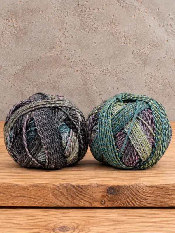 Two round balls of Schoppel-Wolle Edition 3 variegated yarn in shades of green, purple, and charcoal resting on a wooden surface.