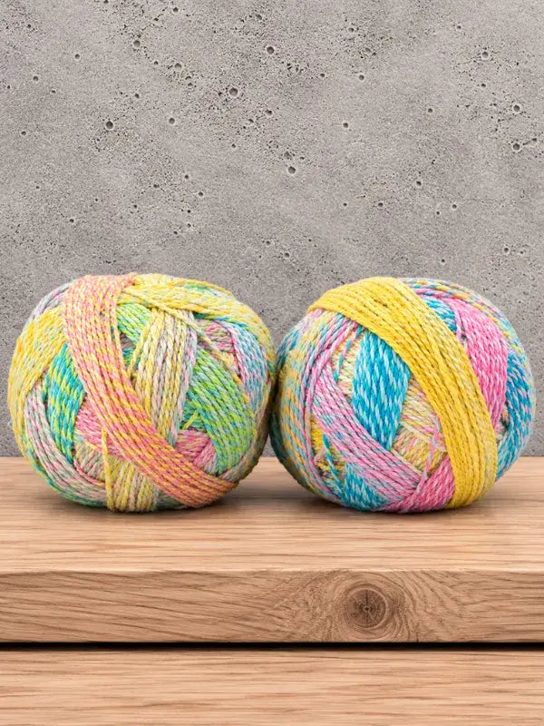 Two balls of Schoppel-Wolle Edition 3 yarn in pastel shades of yellow, pink, blue, and green resting on a wooden surface.