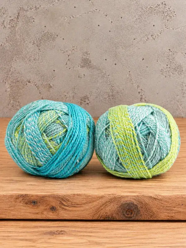Two balls of variegated Schoppel-Wolle Edition 3 yarn in shades of blue and lime green sitting on a wooden surface.