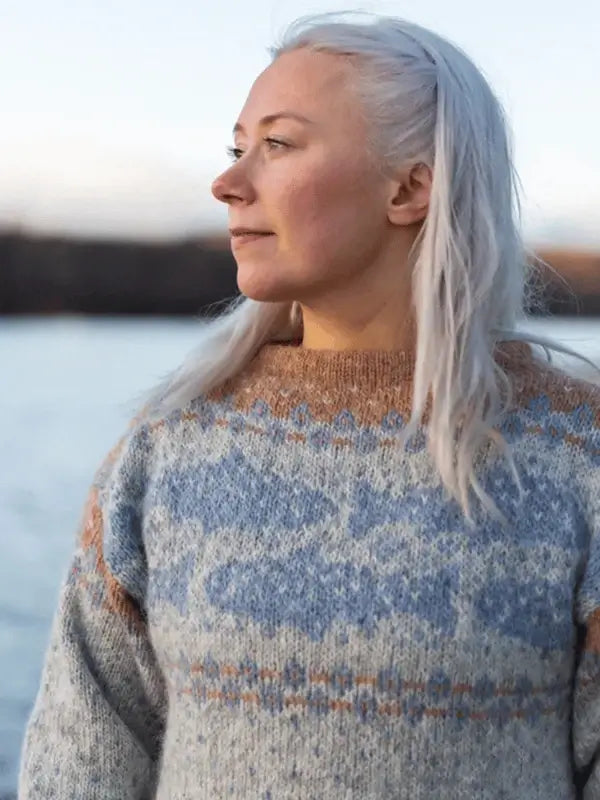 A woman with long, light gray hair wears the Sisu Knitwear Lohineule neuleohje Suomi PDF sweater and stands by the water, gazing thoughtfully into the distance. The softly blurred background highlights the cozy knit feel.