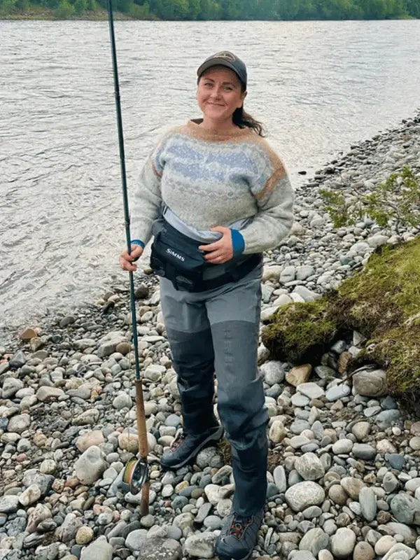 Wearing a Sisu Knitwear Lohineule neuleohje Suomi PDF sweater, waders, boots, and a cap, a person stands on a rocky riverbank holding a fishing rod and smiling at the camera with trees and water in the background.