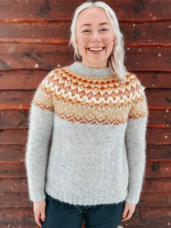 A smiling person with long, light hair wears a Sisu Knitwear Mikkelineule neuleohje Suomi PDF–a light gray sweater with a yellow, orange, and white yoke–standing before a wooden wall as snow falls around them.