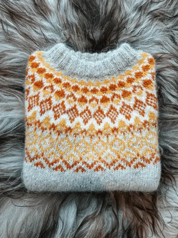 A folded, light gray Sisu Knitwear Mikkelineule neuleohje Suomi PDF sweater with a yellow-orange diamond pattern rests on soft gray fur—ideal as an ulkoilupaita for cozy outings or relaxing after perhokalastus adventures.
