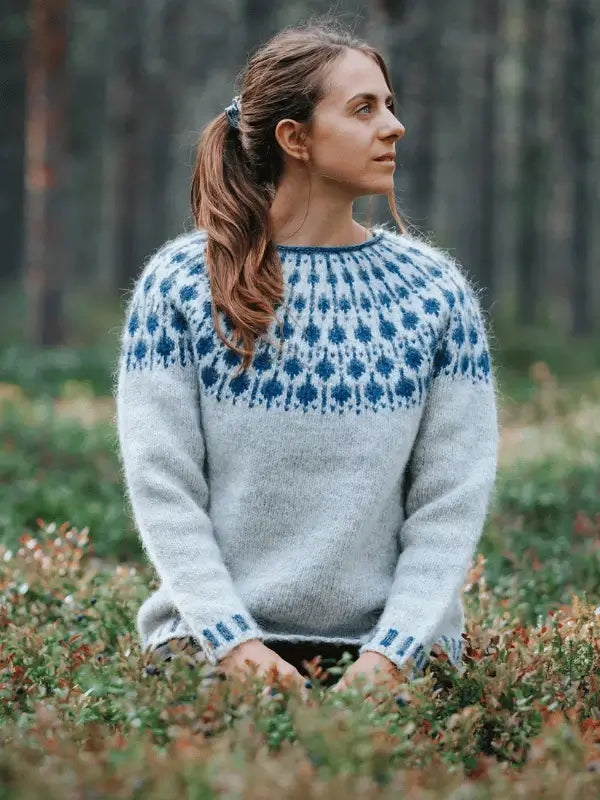 A person with long brown hair in a ponytail sits outdoors in a green forest, wearing a light grey Sisu Knitwear Mustikkaneule sweater made from the Suomi PDF pattern. They are looking to the side.