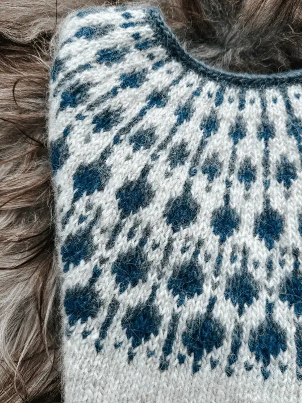 A close-up of the Sisu Knitwear Mustikkaneule neuleohje Suomi PDF pattern shows a round yoke sweater with white, blue, and gray designs, lying on a surface with long, wavy brown hair around it.
