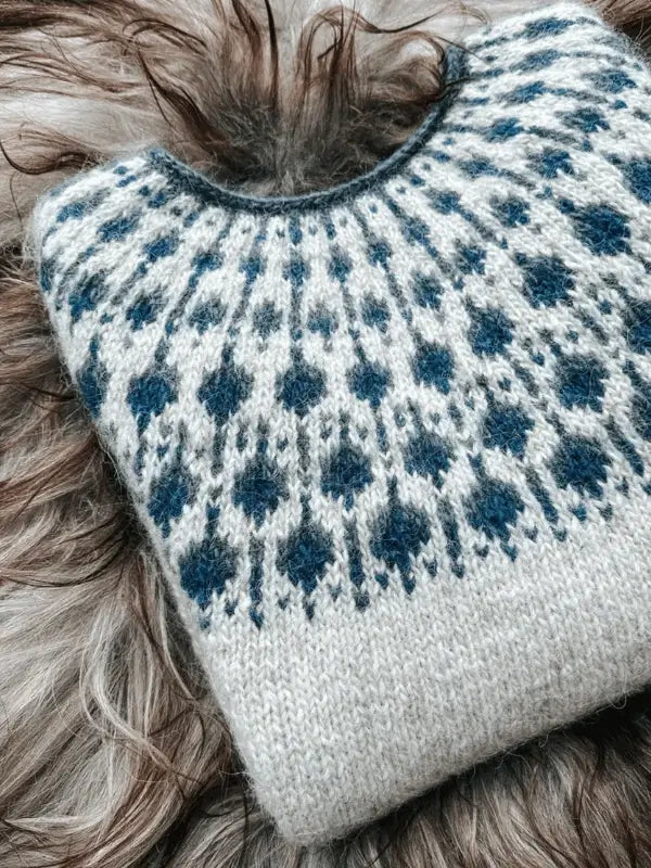 A folded Sisu Knitwear Mustikkaneule neuleohje Suomi PDF sweater with a circular yoke in blue and white rests on soft brown and gray fur.