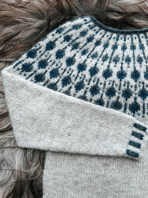 A close-up of a Sisu Knitwear Mustikkaneule neuleohje Suomi PDF sweater with a light gray body and sleeves, plus a blue and dark gray patterned yoke, displayed on a fluffy textured surface.