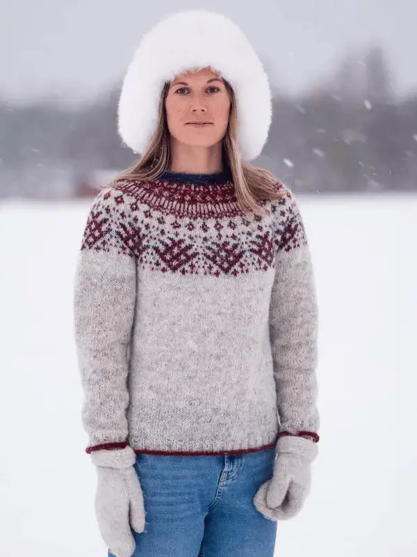 A woman stands outside in falling snow wearing a fluffy white hat, blue jeans, and a gray Rovaneule sweater made using the Sisu Knitwear Rovaneule neuleohje Suomi PDF. The wintry background is gently blurred.