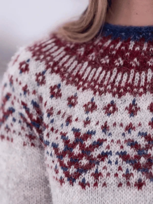 Close-up of a person wearing a Sisu Knitwear Rovaneule neuleohje Suomi PDF wool sweater featuring a traditional Nordic pattern in red, blue, and white; only the lower half of the face and upper torso are visible.