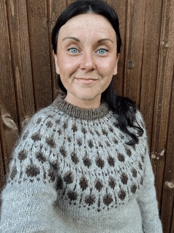 A woman with fair skin, blue eyes, and dark hair smiles at the camera wearing the Sisu Knitwear Terhoneule neuleohje Suomi PDF, featuring a brown pyöreä kaarroke pattern, as she stands before a wooden wall.